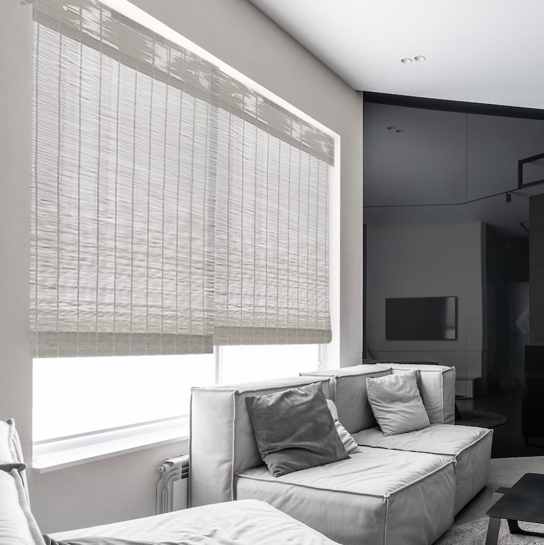 May include: White woven bamboo blinds in a modern living room with a grey sectional sofa and grey throw pillows.