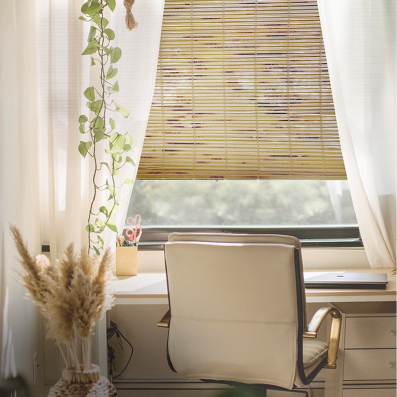 May include: A white office chair with gold accents sits in front of a window with bamboo blinds. The blinds are partially open, allowing natural light to filter into the room. A desk with a laptop and a vase of dried flowers is visible in the background.