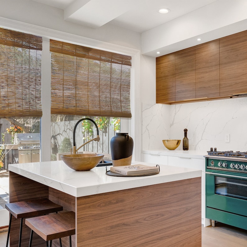 May include: A modern kitchen island with a white worktop and a wooden base. The island has a large sink with a gold tap, a bowl, and a book on top. The kitchen has a green stove and a white worktop with a gold bowl.