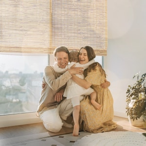 May include: A family of three, a man, a woman, and a young child, are hugging each other. The woman is wearing a yellow floral dress, the man is wearing a light brown shirt, and the child is wearing a white dress. They are standing in front of a window with bamboo blinds.