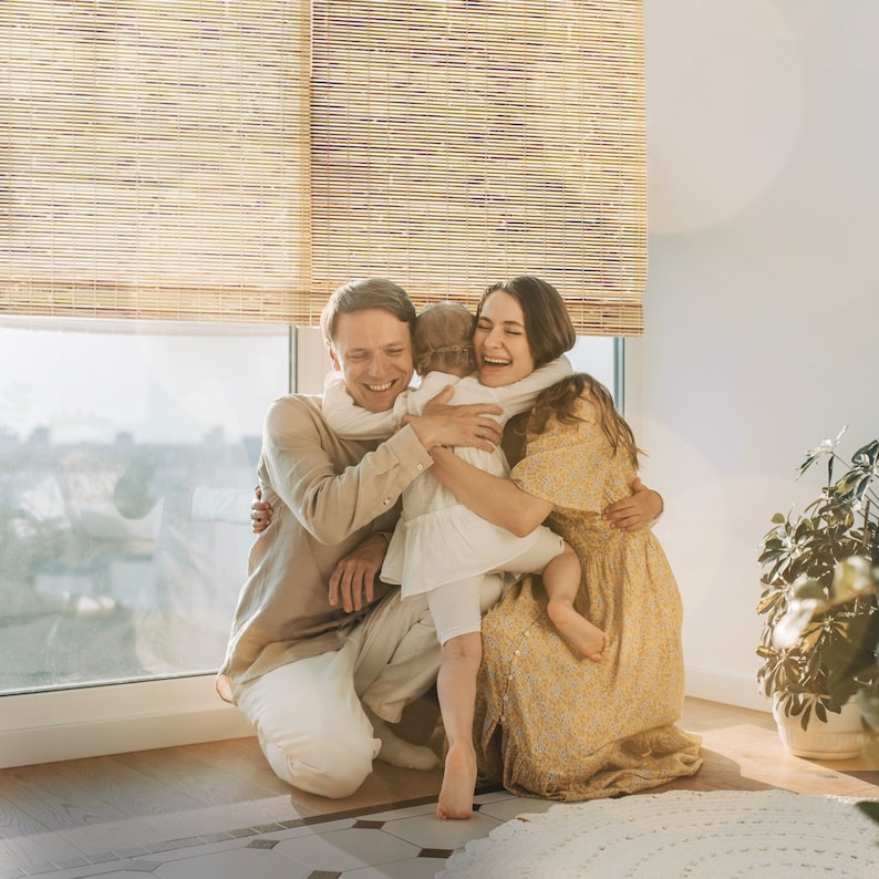 May include: A family of three, a man, a woman, and a young child, are hugging each other. The woman is wearing a yellow floral dress, the man is wearing a light brown shirt, and the child is wearing a white dress. They are standing in front of a window with bamboo blinds.