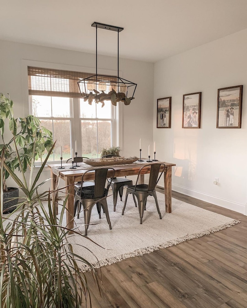 May include: A rustic wooden dining table with black metal chairs, a white rug, and a black metal chandelier with a leafy design. The table is set with a wooden bowl, candles, and a plant.
