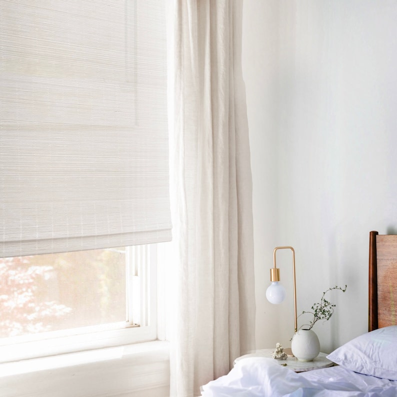 May include: A white bamboo roman blind is partially lowered in a bedroom window. The blind is paired with a white sheer curtain. A bedside table with a gold lamp and a vase of white flowers is next to a bed with white bedding.