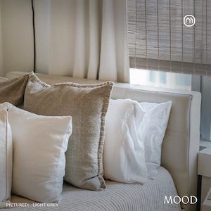 May include: A white leather headboard with three pillows. The pillows are white, beige, and light grey. The headboard is against a white wall with a window behind it. The window has a light grey woven blind pulled down. The text "MOOD" is in the lower right corner of the image. The text "PICTURED: LIGHT GREY" is in the lower left corner of the image.