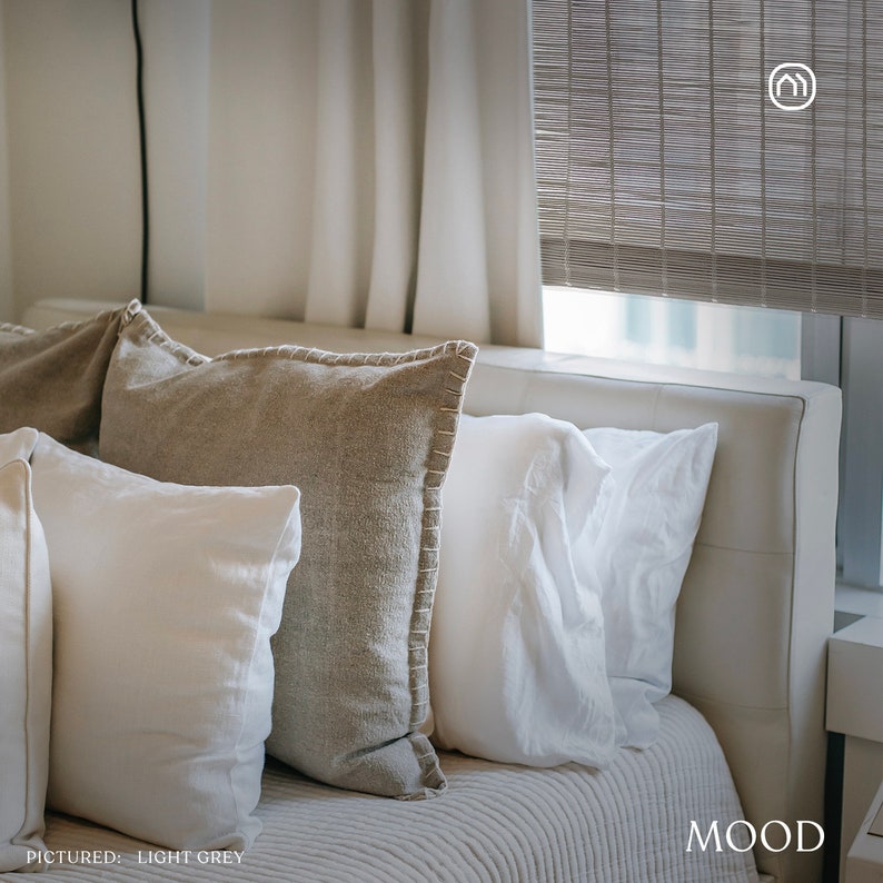May include: A white leather headboard with three pillows. The pillows are white, beige, and light grey. The headboard is against a white wall with a window behind it. The window has a light grey woven blind pulled down. The text "MOOD" is in the lower right corner of the image. The text "PICTURED: LIGHT GREY" is in the lower left corner of the image.