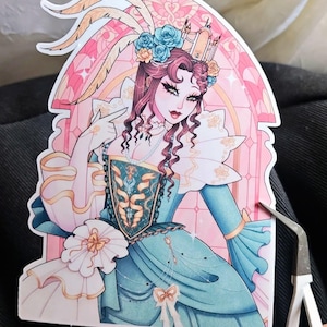 Puede incluir: Una pegatina con una ilustraci&oacute;n estilizada de una mujer con un vestido turquesa y blanco, adornada con un tocado de plumas y detalles florales. La obra de arte tiene un fondo rosa y dorado.
