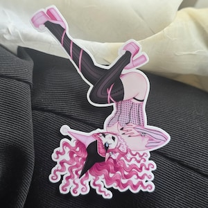 May include: A die-cut sticker featuring a stylized character with long, wavy pink hair, a black and pink jester hat, and a black and pink corset. The character wears black striped stockings and pink platform shoes, posed in a dynamic, inverted position.