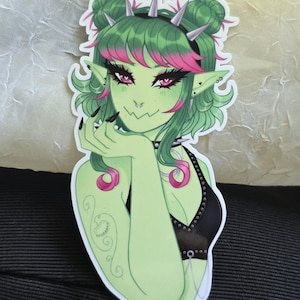 May include: A sticker featuring a green-skinned character with green hair, pink highlights, and pink eyes. The character wears a black top and a spiked headband. A tattoo is visible on the character's arm. The character is smiling and touching their face.