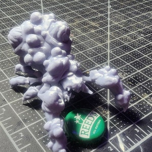 May include: A light purple, 3D-printed creature with multiple limbs and a bulbous body. A green and white Reed's All Natural bottle cap is next to the creature. The creature is on a black and gray grid surface.