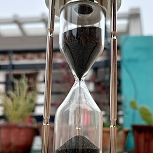 Pode incluir: Um relógio de areia prateado com areia preta, em pé sobre uma superfície branca. O relógio de areia é feito de metal e vidro, e tem um design clássico.