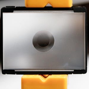 May include: A rectangular, silver-toned object with a dark circular indentation in the center. The object is framed in black and supported by two yellow, rectangular blocks. The background is blurred.