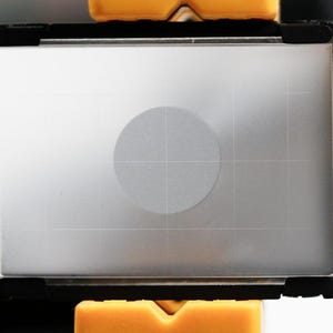 May include: A rectangular, silver-toned camera accessory with a black frame and a circular detail in the center. The accessory has a grid pattern and is held by two yellow supports.