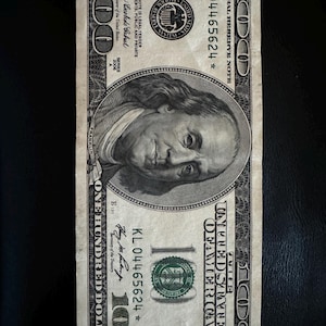May include: A US $100 bill with a portrait of Benjamin Franklin. The bill has the serial number KL 04465624 and the text "UNITED STATES OF AMERICA" and "ONE HUNDRED DOLLARS".