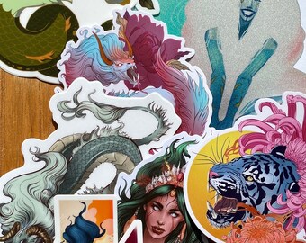 May include: A collection of colorful stickers featuring various mythical creatures, including a dragon, a seahorse, a tiger, and a woman with a crown. The stickers are on a wooden surface.