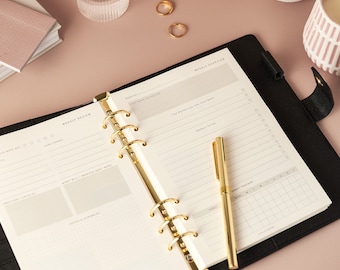 May include: An open black planner with gold accents, featuring a weekly overview and review layout. A gold pen rests on the page. The planner is on a pink surface, with a glass of water, a small notebook, and two gold rings.