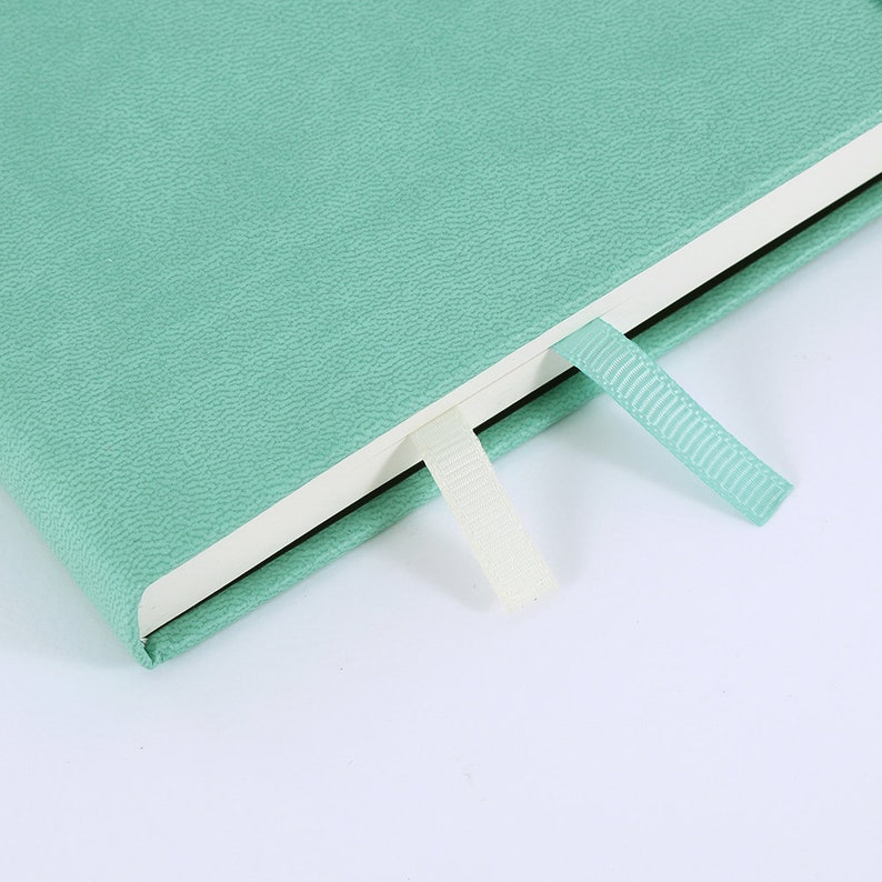K&ouml;nnte beinhalten: Nahaufnahme eines mintgr&uuml;nen Journals mit strukturierter H&uuml;lle. Das Journal ist ge&ouml;ffnet und zeigt wei&szlig;e Seiten und zwei Leseb&auml;ndchen, eines cremefarbenes und eines mintgr&uuml;nes. Der Einband des Journals hat eine subtile, genarbte Textur.