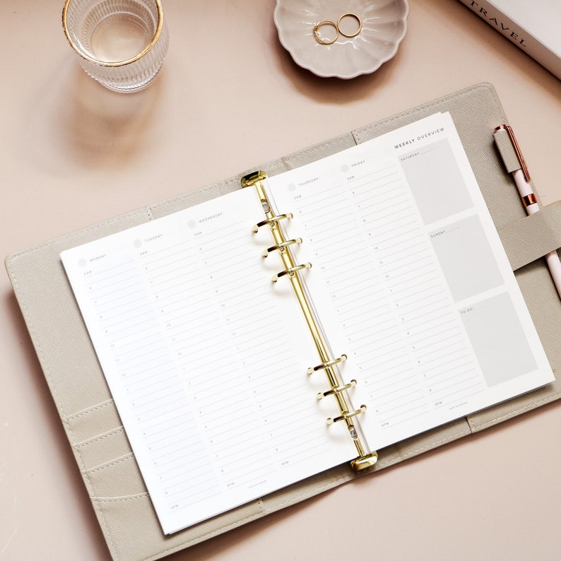 May include: Open gray planner with gold accents, featuring a weekly overview layout. The planner is open to a blank page with a detailed schedule. A pen, rings, and a glass are also visible.