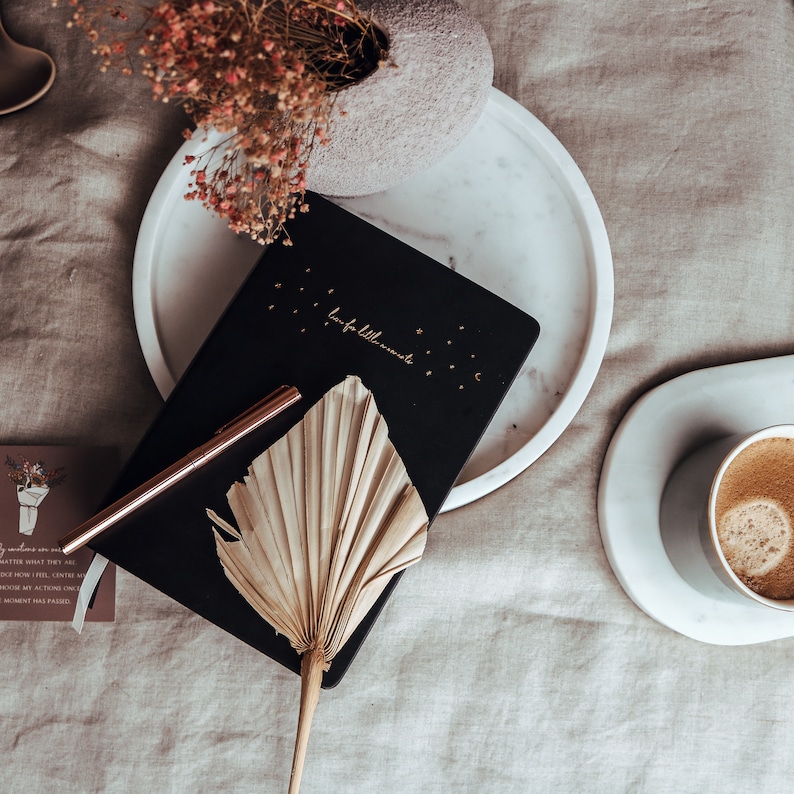 K&ouml;nnte beinhalten: Draufsicht mit einem schwarzen Notizbuch mit goldener Schrift, einem ros&eacute;goldenen Stift und einem getrockneten Palmblatt. Eine Tasse Kaffee, eine dekorative Vase und eine kleine Karte sind ebenfalls vorhanden. Das Notizbuch tr&auml;gt den Schriftzug "love for little moments".