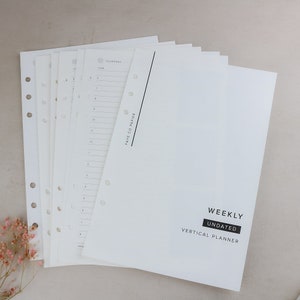 May include: A stack of white undated weekly vertical planner pages. The pages are pre-punched with holes for binding. The cover page has the words "WEEKLY UNDATED VERTICAL PLANNER" in a black rectangle.