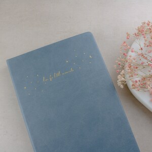 K&ouml;nnte beinhalten: Ein blaues Notizbuch mit goldenen Sternen, einem Mond und dem Schriftzug "love for little moments". Das Notizbuch liegt auf einer hellen Oberfl&auml;che, mit einem wei&szlig;en Teller und rosa Blumen im Hintergrund.