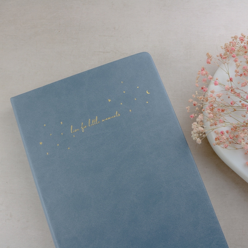 K&ouml;nnte beinhalten: Ein blaues Notizbuch mit goldenen Sternen, einem Mond und dem Schriftzug "love for little moments". Das Notizbuch liegt auf einer hellen Oberfl&auml;che, mit einem wei&szlig;en Teller und rosa Blumen im Hintergrund.