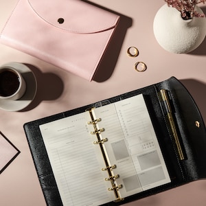 May include: A black leather planner with gold accents, open to a daily planning page. A pink leather envelope, a cup of coffee, and two gold rings are arranged on a pink surface. A small white vase with dried flowers is also present.