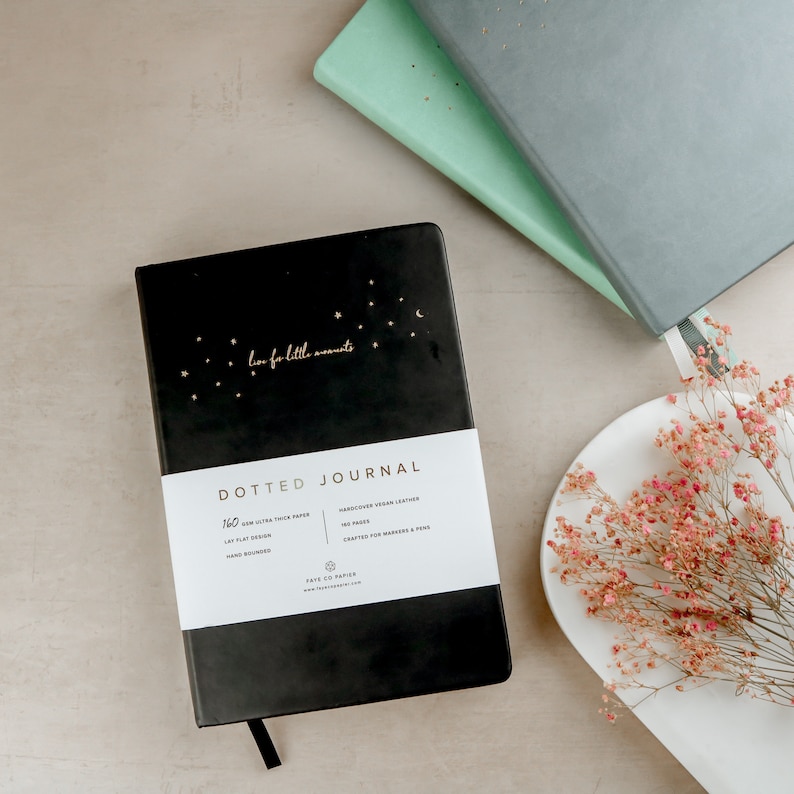 K&ouml;nnte beinhalten: Ein schwarzes Notizbuch mit Punkten, goldenen Sternen und dem Schriftzug "live for little moments". Das Notizbuch hat ein wei&szlig;es Band mit Produktdetails. Zwei weitere Notizb&uuml;cher in Grau und Mintgr&uuml;n sind sichtbar. Eine wei&szlig;e Platte mit rosa Blumen ist ebenfalls im Bild.