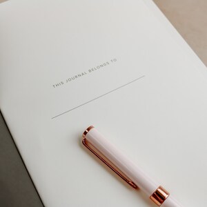 K&ouml;nnte beinhalten: Ein Notizbuch mit dem Text "THIS JOURNAL BELONGS TO" auf dem Einband. Ein hellrosa Stift mit goldfarbenen Akzenten liegt auf dem Notizbuch. Das Notizbuch ist auf einer leeren Seite ge&ouml;ffnet.