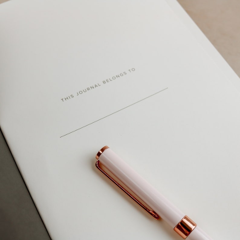 K&ouml;nnte beinhalten: Ein Notizbuch mit dem Text "THIS JOURNAL BELONGS TO" auf dem Einband. Ein hellrosa Stift mit goldfarbenen Akzenten liegt auf dem Notizbuch. Das Notizbuch ist auf einer leeren Seite ge&ouml;ffnet.