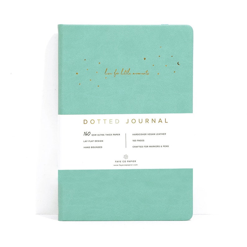K&ouml;nnte beinhalten: Ein t&uuml;rkisblaues Notizbuch mit Punkten, dem Text "love for little moments" und goldenen Sternen und einem Mond. Das Notizbuch hat ein wei&szlig;es Band mit dem Text "DOTTED JOURNAL" und Details zu Papier und Einband.