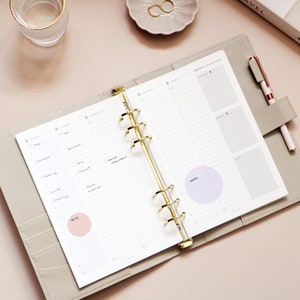 May include: Open weekly planner with gold rings, featuring a light gray cover and lined pages. The planner is open to a weekly overview with handwritten notes and two colored circles for notes. A pen rests in the planner's holder.