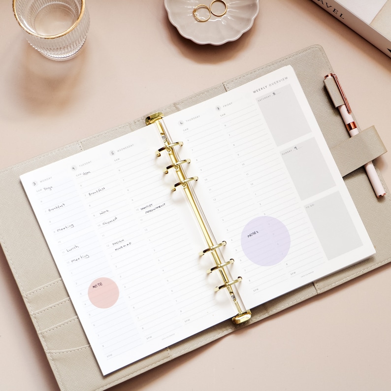 May include: Open weekly planner with gold rings, featuring a light gray cover and lined pages. The planner is open to a weekly overview with handwritten notes and two colored circles for notes. A pen rests in the planner's holder.