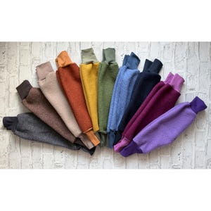 Op de afbeelding: Een verzameling fleece armwarmers in verschillende kleuren, waaronder grijs, bruin, beige, oranje, geel, groen, blauw, zwart, bordeauxrood en paars. Elke armwarmer heeft een contrasterende manchet en kraag, gerangschikt in een waaier.
