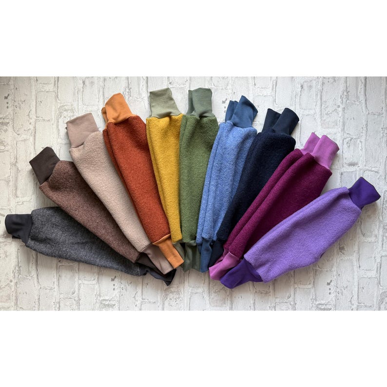 K&ouml;nnte beinhalten: Eine Sammlung von Fleece-Armstulpen in verschiedenen Farben, darunter Grau, Braun, Beige, Orange, Gelb, Gr&uuml;n, Blau, Schwarz, Burgunderrot und Lila. Jede Armstulpe hat einen kontrastierenden Bund und Kragen, die f&auml;cherf&ouml;rmig angeordnet sind.