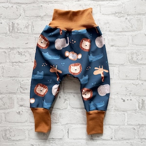 Könnte beinhalten: Babyhose mit einem sich wiederholenden Muster aus Löwen, Elefanten, Affen, Krokodilen und Giraffen auf marineblauem Hintergrund. Die Hose hat einen braunen Bund und Bündchen.