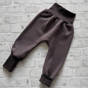 May include: Dark brown waffle knit pants featuring a high, solid-colored elastic waistband and matching dark brown ribbed cuffs at the ankles. The garment has a relaxed fit with a dropped crotch design and a textured, grid-like fabric pattern.