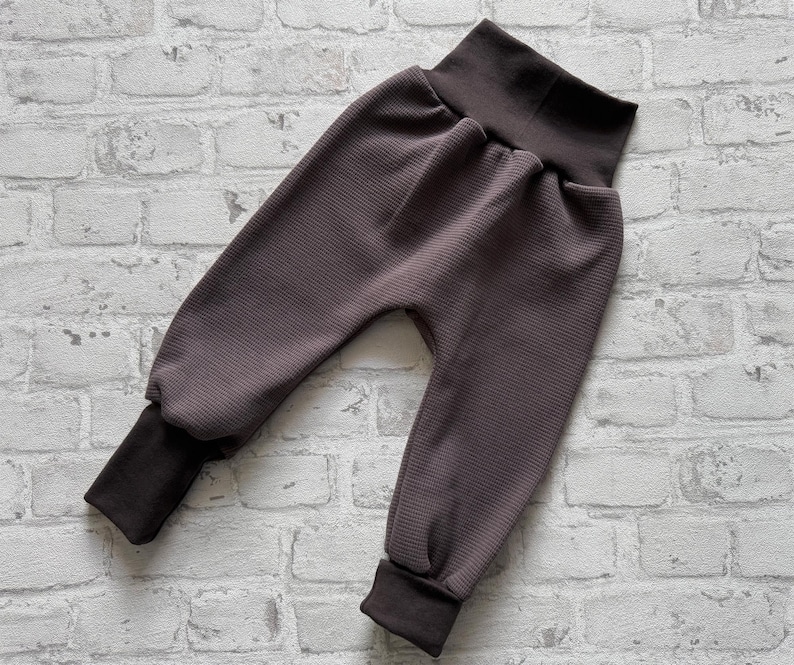 May include: Dark brown waffle knit pants featuring a high, solid-colored elastic waistband and matching dark brown ribbed cuffs at the ankles. The garment has a relaxed fit with a dropped crotch design and a textured, grid-like fabric pattern.