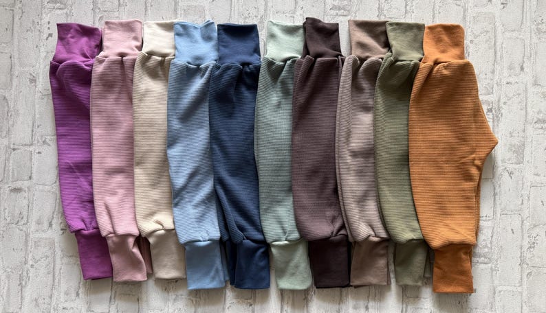 May include: A collection of ten pairs of ribbed knit pants arranged side-by-side in a spectrum of colors including purple, light pink, cream, light blue, navy, sage green, dark brown, taupe, olive green, and mustard yellow with cuffed ankles.