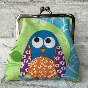 May include: A green, blue, orange, and purple fabric coin purse with a metal clasp. The purse features a cartoon owl with large eyes and a floral pattern.
