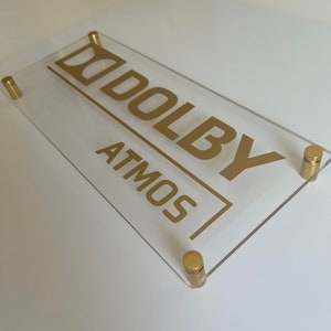 May include: A clear acrylic sign with gold lettering that reads "Dolby Atmos". The sign is mounted with four gold screws.