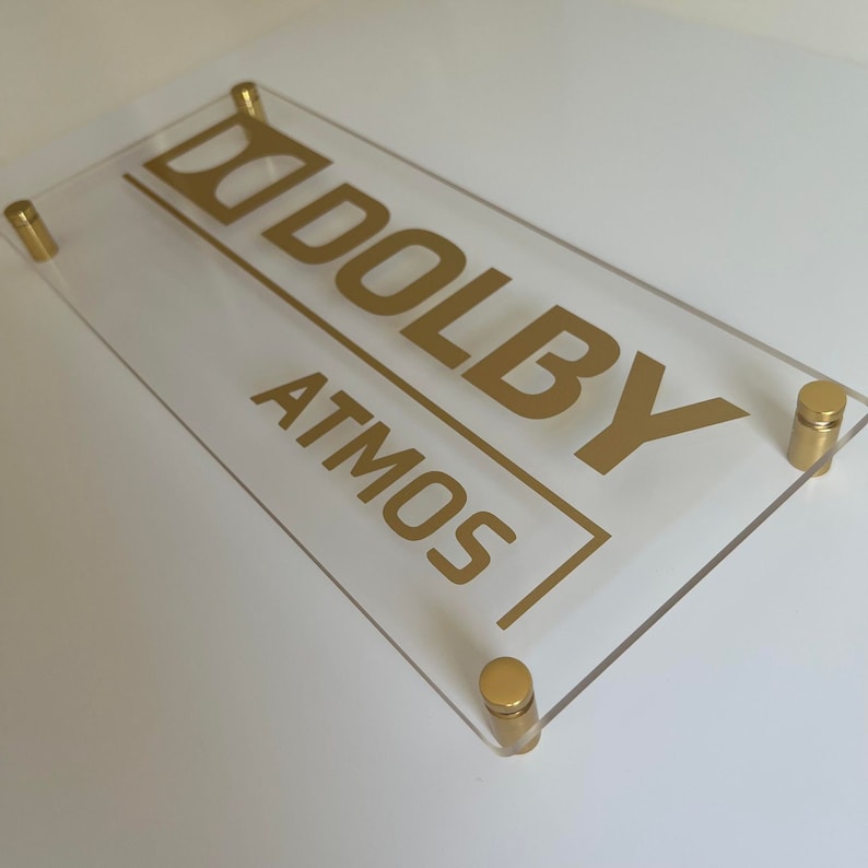 May include: A clear acrylic sign with gold lettering that reads "Dolby Atmos". The sign is mounted with four gold screws.
