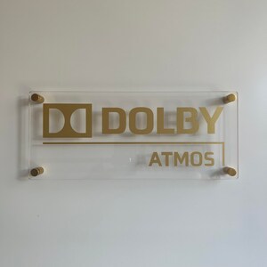 May include: A clear acrylic sign with gold lettering that reads "Dolby Atmos". The sign is mounted with gold screws.