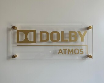 May include: A clear acrylic sign with gold lettering that reads "Dolby Atmos". The sign is mounted with gold screws.