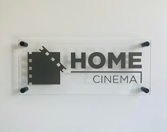 May include: A clear acrylic sign with black lettering that reads "HOME CINEMA". A stylized film reel icon is to the left of the text.