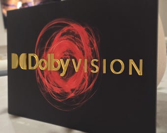 May include: A black rectangular sign with the Dolby Vision logo. The logo features gold lettering and a red, swirling design. The sign is displayed on a light-colored surface.