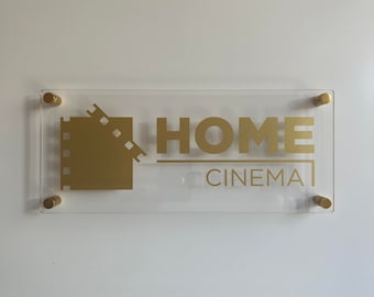 May include: A clear acrylic sign with gold lettering that reads "HOME CINEMA". A gold film reel icon is to the left of the text.