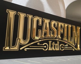May include: A black rectangular sign with the gold text "LUCASFILM Ltd". The letters are raised and have a metallic, reflective appearance. The sign is angled, showing the logo's design.