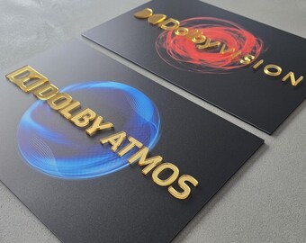 May include: Two black cards with gold lettering and logos. One card features the words "DOLBY ATMOS" with a blue and white graphic. The other card has "DOLBY VISION" with a red and orange graphic. The cards are on a gray surface.