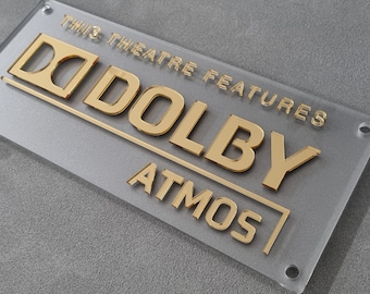 May include: Clear acrylic sign with gold lettering that reads "THIS THEATRE FEATURES DOLBY ATMOS." The sign has four corner holes and is accompanied by gold and black mounting hardware. The sign is on a gray surface.