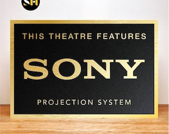 May include: A black rectangular sign with gold lettering and a gold border. The text reads "THIS THEATRE FEATURES SONY PROJECTION SYSTEM". The sign is on a wooden surface.